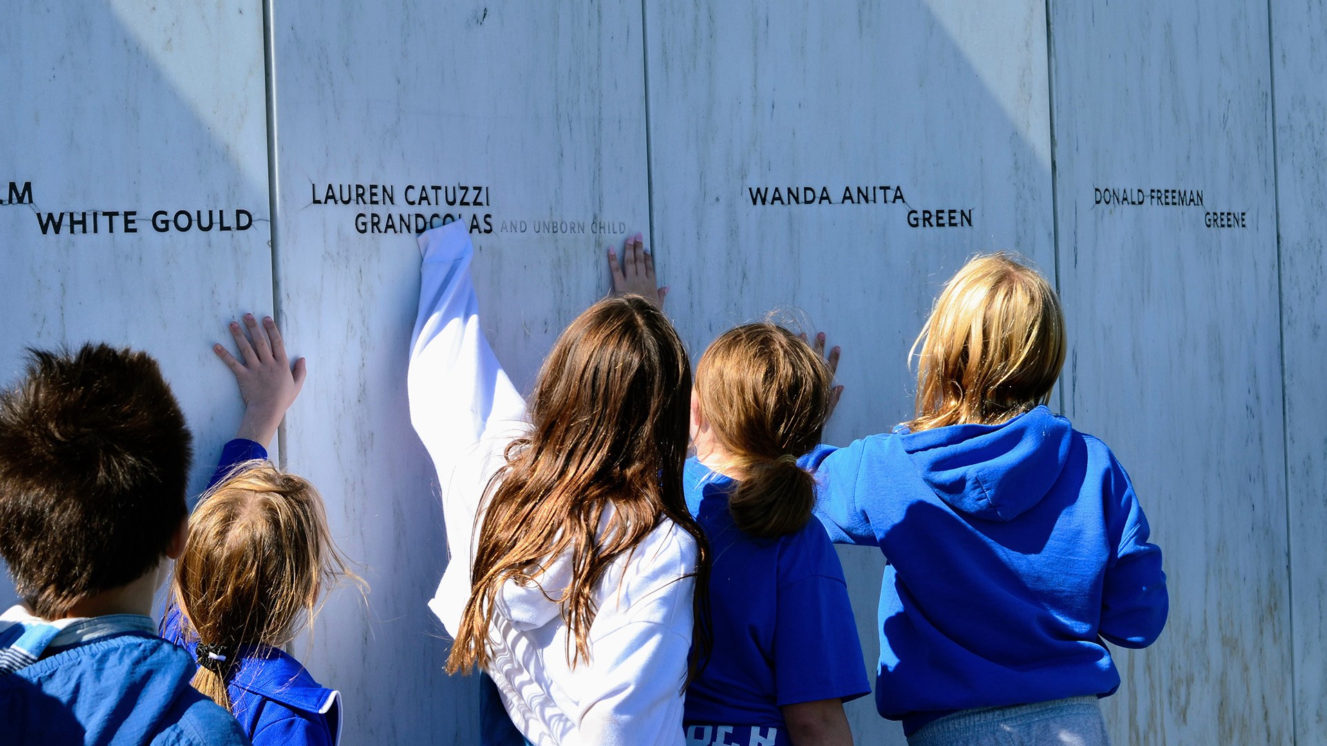 Students touching the engraved names of the forty passengers and crew members of Flight 93 at a white marble wall.